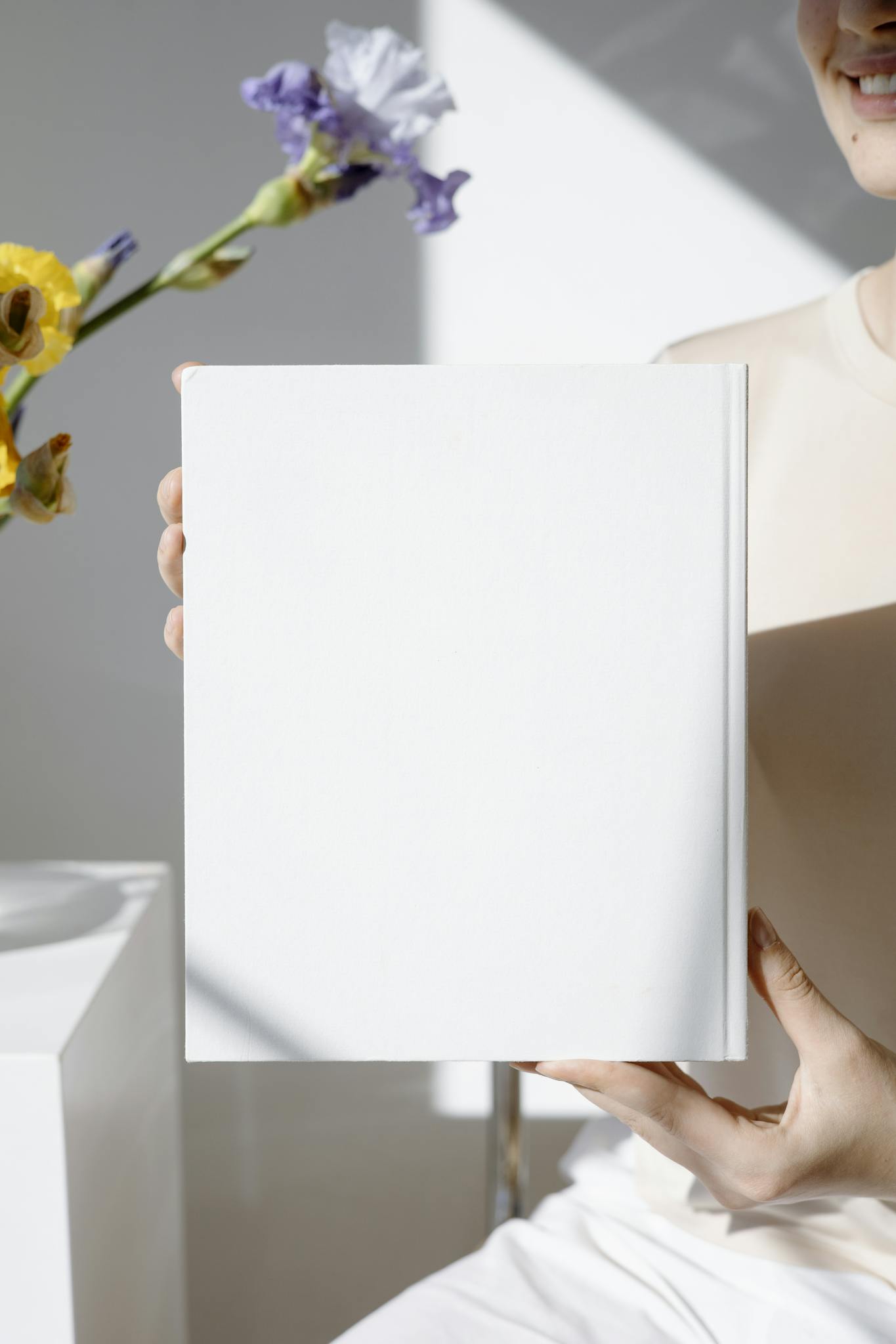 Minimalist blank book cover held in a bright room with flowers in the background.